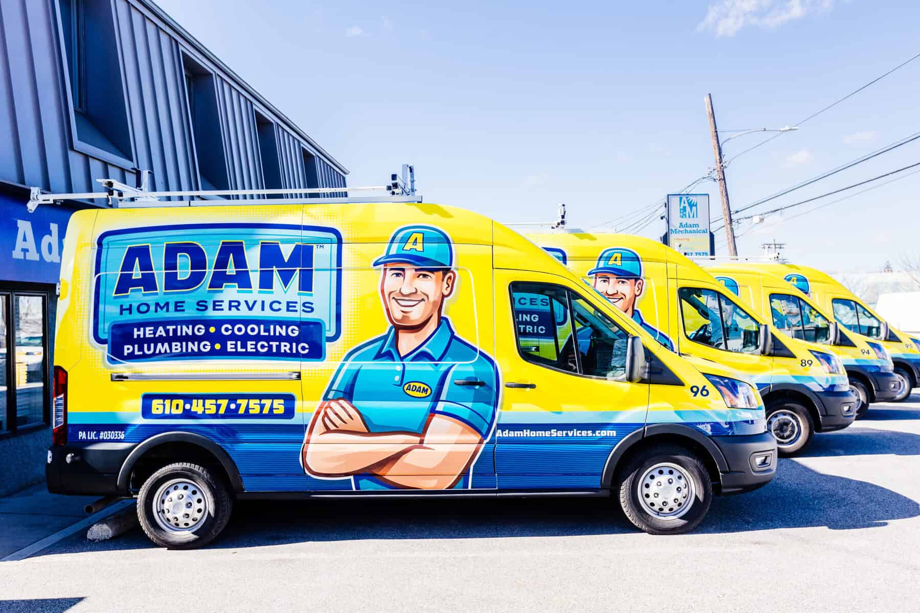 Yellow service vans with branding for "Adam Home Services" parked in a row outside a building. The vans, featuring a cartoon technician, list services like heating, cooling, plumbing, electric, and drain clearing service.