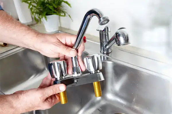 Person installing or removing a silver kitchen faucet over a stainless steel sink, with a plant in the background.