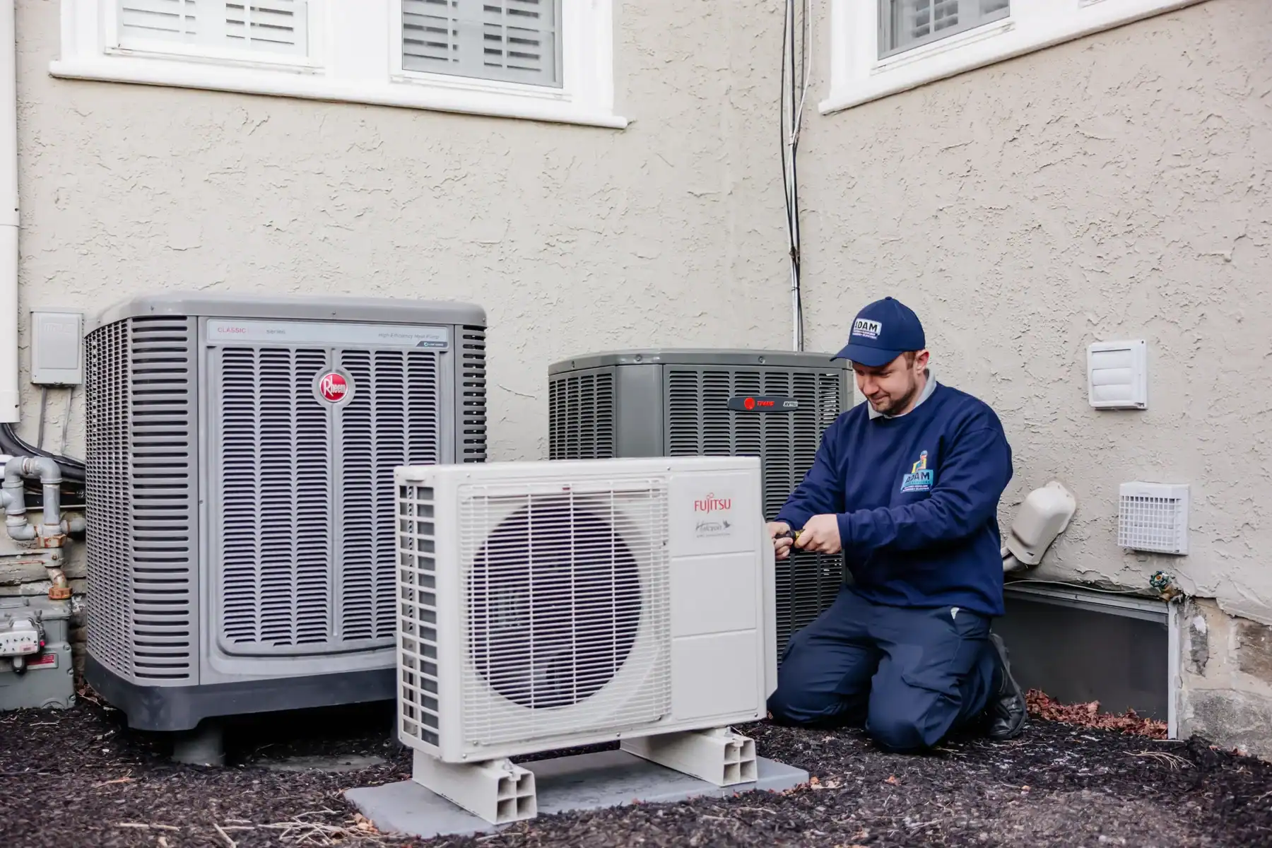 A technician in a uniform kneels beside an outdoor air conditioning unit, using a tool, with two other units nearby against a building wall.