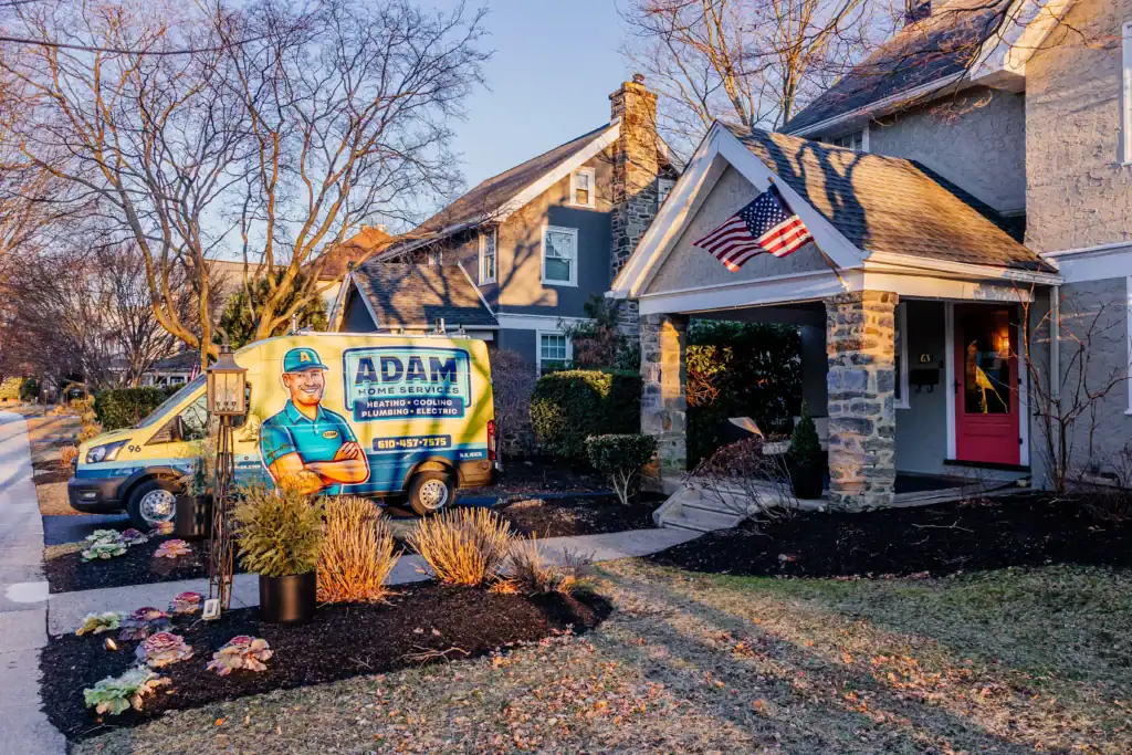 A plumbing service van is parked in front of a house with an American flag and a red door on a suburban street lined with other homes.