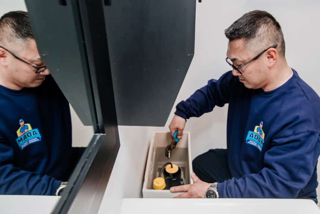 A person in a blue shirt uses pliers to adjust plumbing inside a toilet tank, reflected in a nearby mirror—demonstrating hands-on drain services expertise.