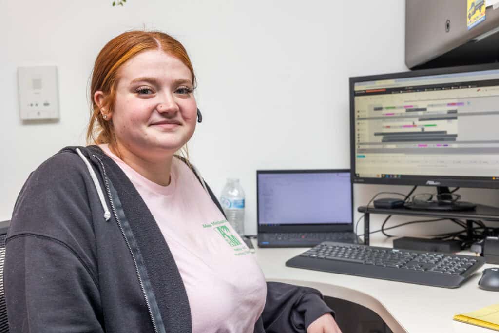 A woman with red hair sits at a desk in an office, facing the camera. Behind her, a computer monitor displays a scheduling application, possibly arranging appointments for heating services or emergency heating repair.