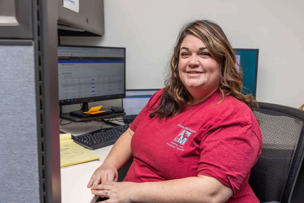 A woman in a red shirt sits at a desk with dual monitors, paperwork, and a keyboard in an office setting, smiling at the camera as she schedules heating services and furnace repair appointments.