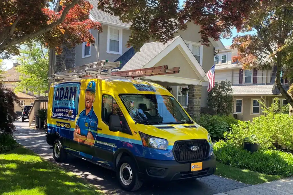 A blue and yellow Adam Services van with roof ladders is parked in the driveway of a suburban house with an American flag.