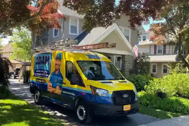 A brightly colored service van with a ladder on top is parked in the driveway of a suburban house on a sunny day.